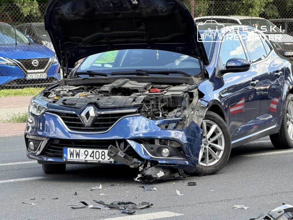 Zniszczony niebieski samochód osobowy marki Renault stoi na drodze, z otwartą maską i uszkodzoną przednią częścią.