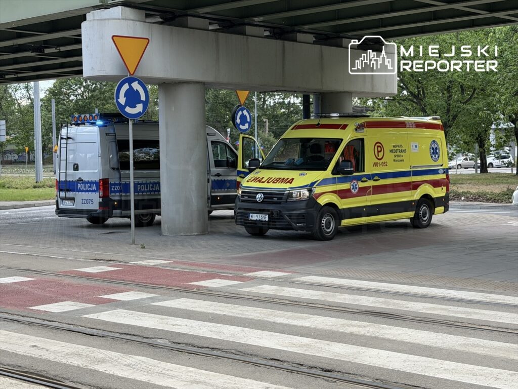 Karetka pogotowia i radiowóz policyjny stoją na skrzyżowaniu pod mostem, w otoczeniu znaków drogowych.