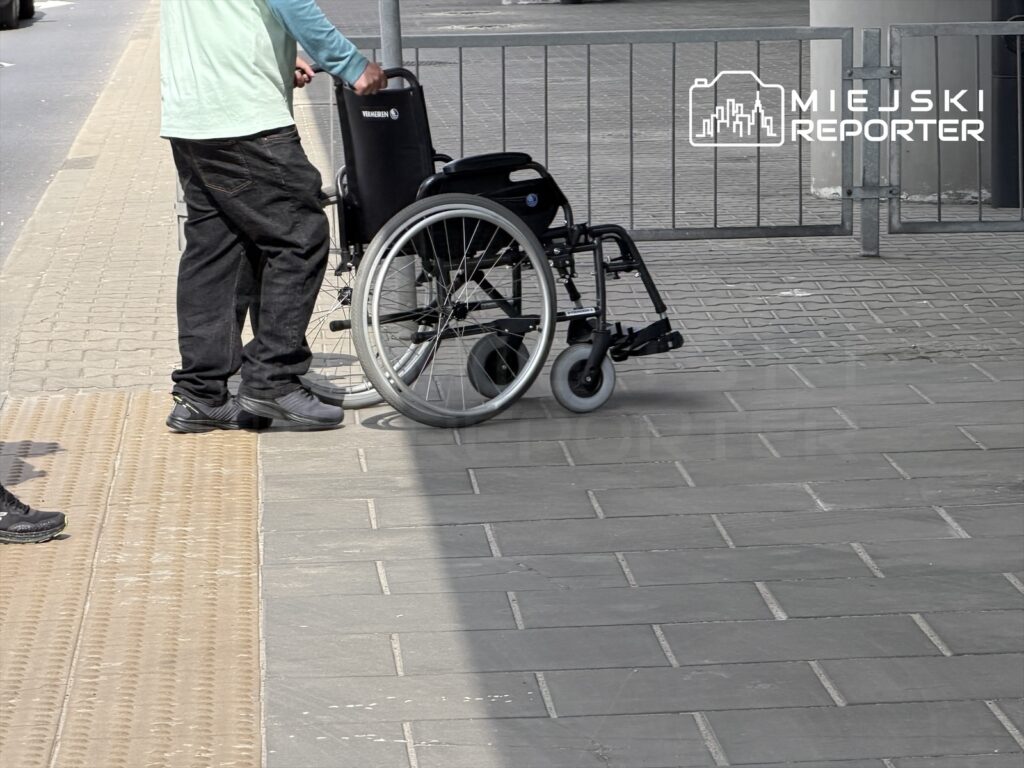 Mężczyzna w niebieskiej koszuli trzyma wózek inwalidzki na chodniku obok przejścia dla pieszych.