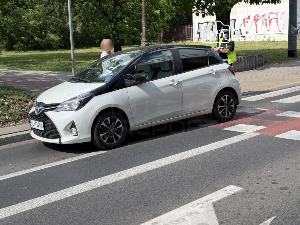 Biały samochód osobowy zatrzymany na przejściu dla pieszych, obok stoi mężczyzna w kamizelce odblaskowej.