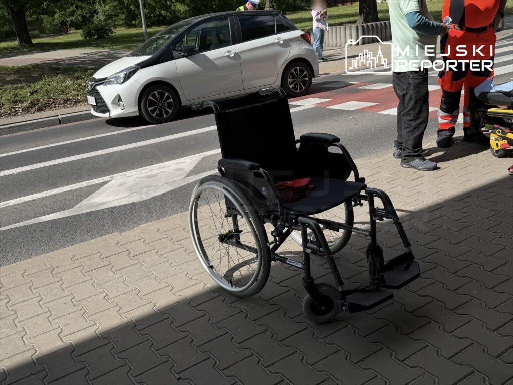 Wózek inwalidzki stoi na chodniku obok zespołu ratownictwa medycznego oraz zaparkowanego samochodu.