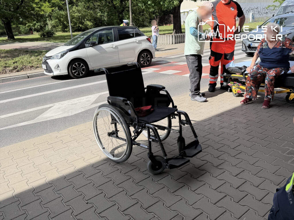 Na chodniku stoi wózek inwalidzki, obok grupka osób, w tym ratownik medyczny, rozmawiająca z kobietą siedzącą na krawężniku.
