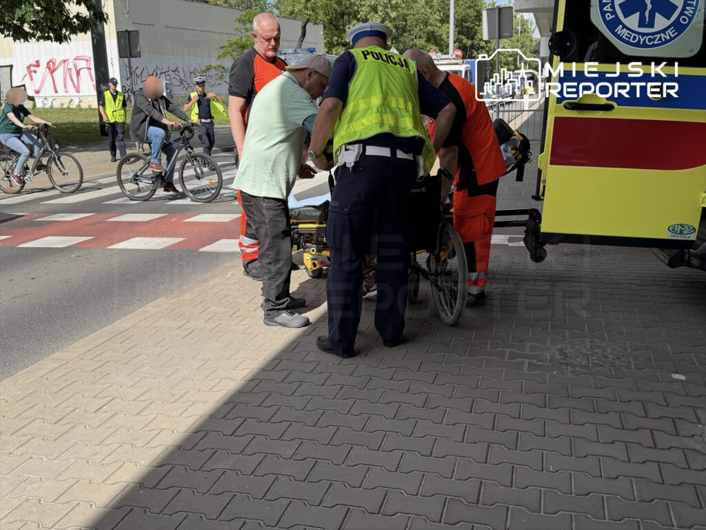 Grupa ratowników medycznych oraz policjantów udziela pomocy osobie na wózku inwalidzkim obok karetki.