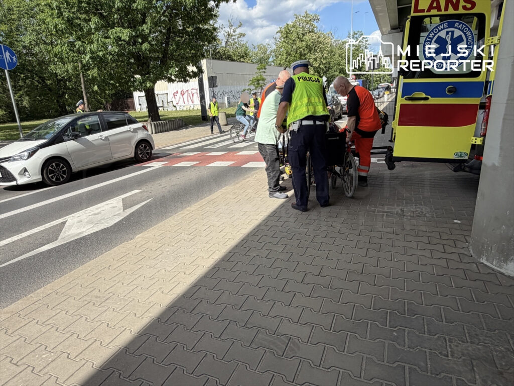 Ratownicy medyczni oraz policja udzielają pomocy poszkodowanemu na skrzyżowaniu, obok karetki.