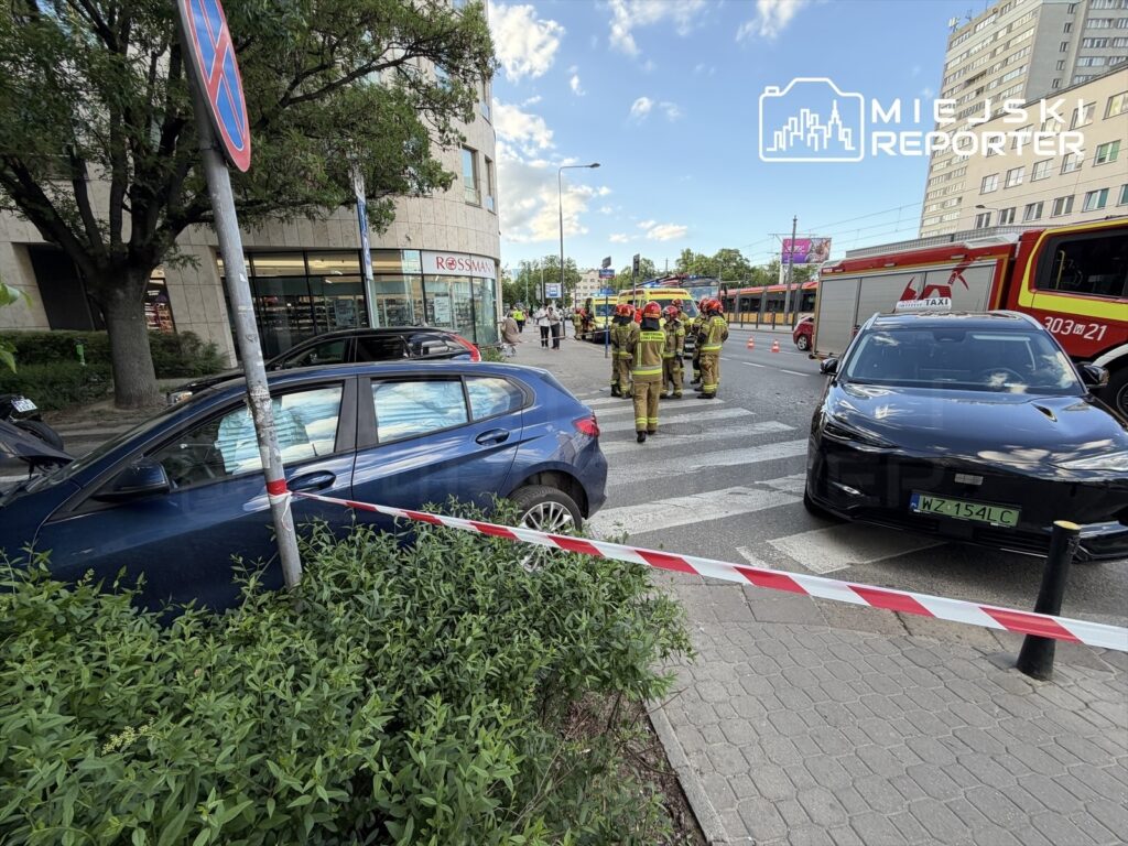 Grupa strażaków w hełmach omawia sytuację na chodniku obok sklepu, podczas gdy samochody stoją w pobliżu.