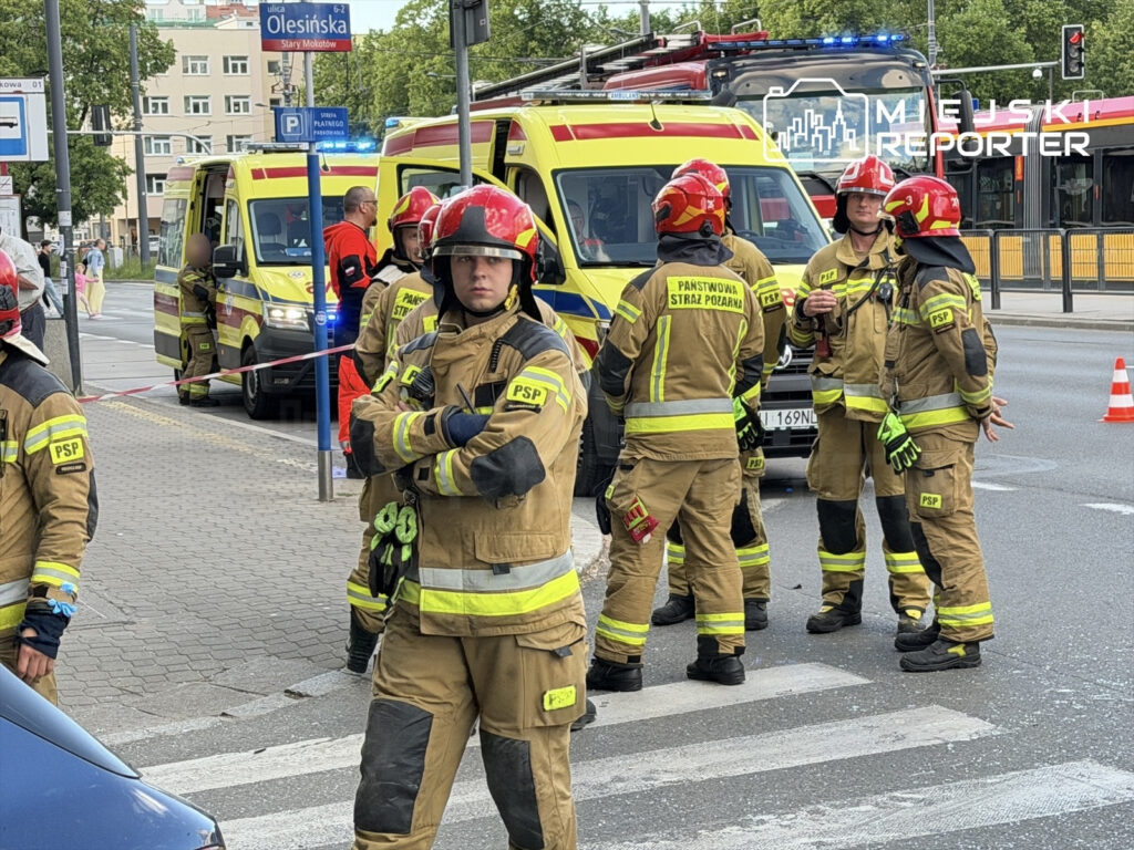 Grupa strażaków w mundurach rozmawia w pobliżu pojazdów ratunkowych na miejskiej ulicy.