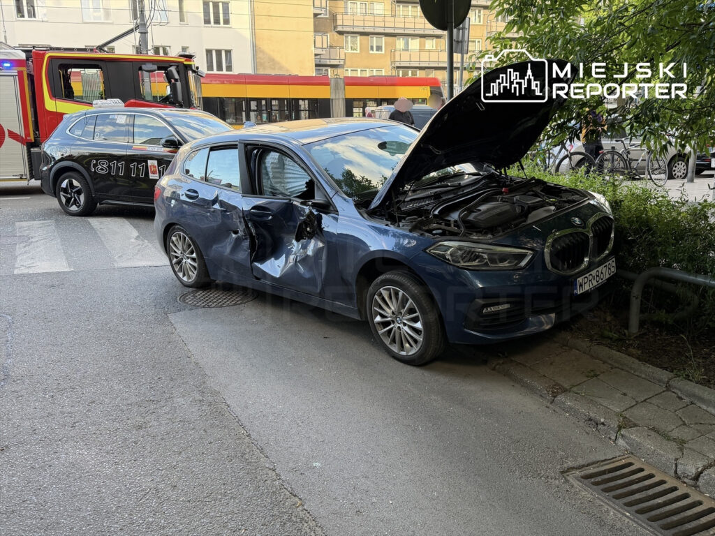 Uszkodzony niebieski samochód osobowy stoi na poboczu, z uniesioną maską; w tle widać pojazdy służb ratunkowych.