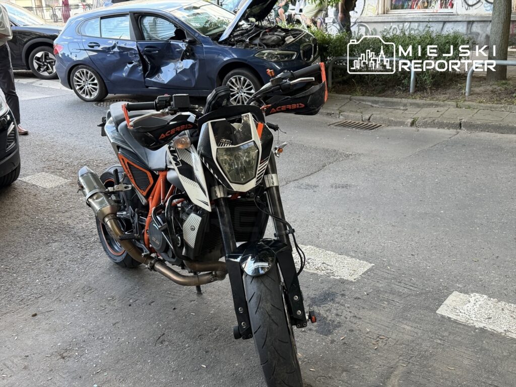 Motocykl sportowy z widocznymi detalami w pomarańczowo-czarnym malowaniu stoi na ulicy obok uszkodzonego samochodu.