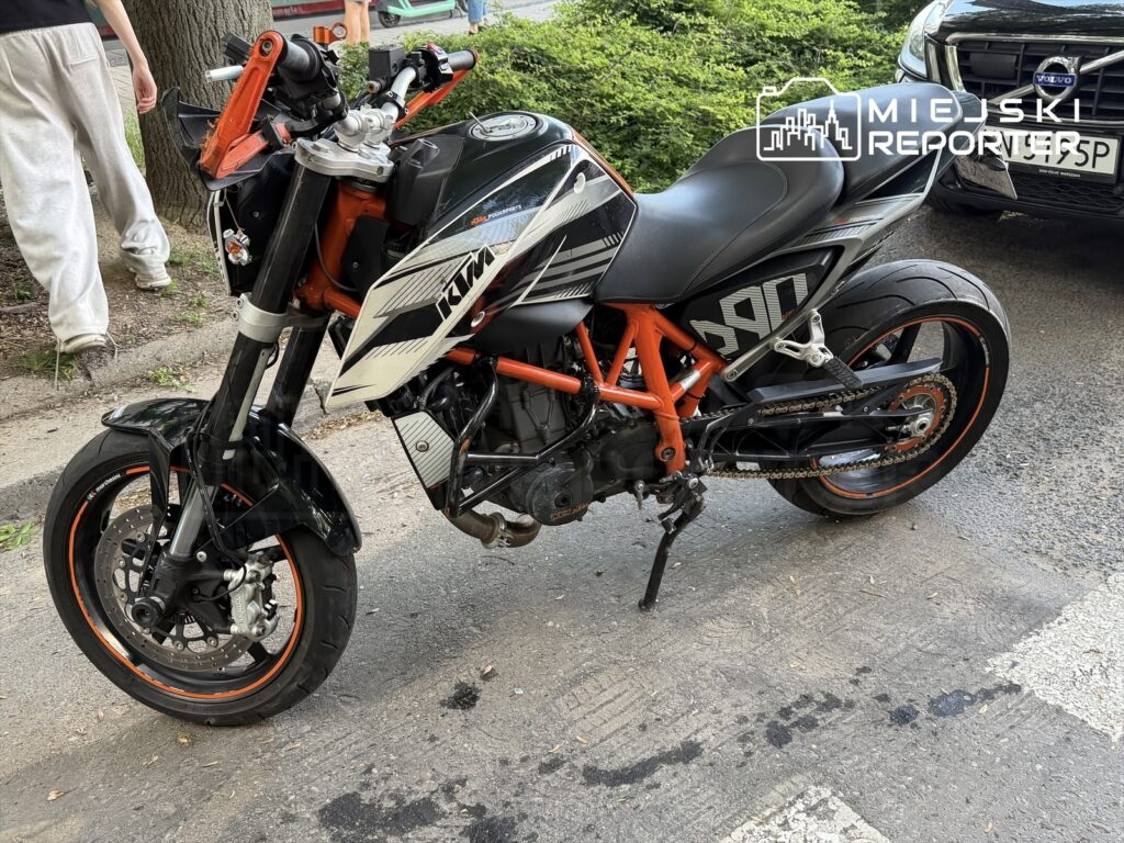 Motocykl marki KTM w czarno-pomarańczowym kolorze zaparkowany na ulicy, obok rosnącego drzewa.