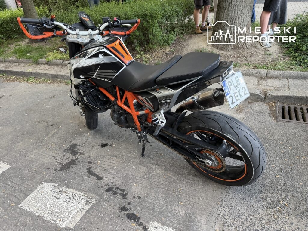 Motocykl sportowy z pomarańczowymi akcentami stoi na ulicy, obok widać zarys ludzi w tle.
