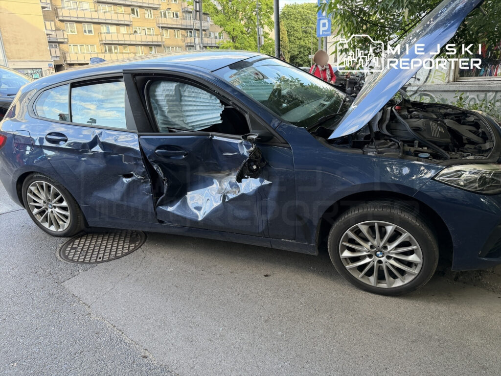 Uszkodzony niebieski samochód osobowy z wgniecionymi drzwiami stoi na ulicy, z otwartą maską silnika.
