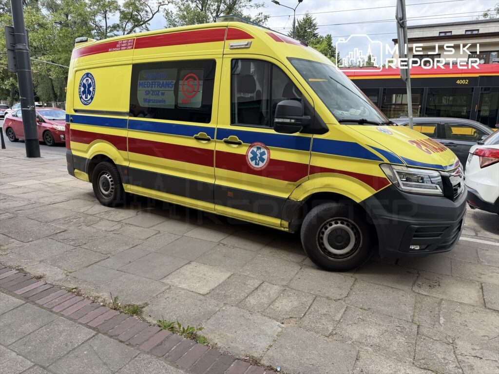 Żółto-czerwony ambulans zaparkowany na ulicy, obok zaparkowanych samochodów, w miejskim otoczeniu.