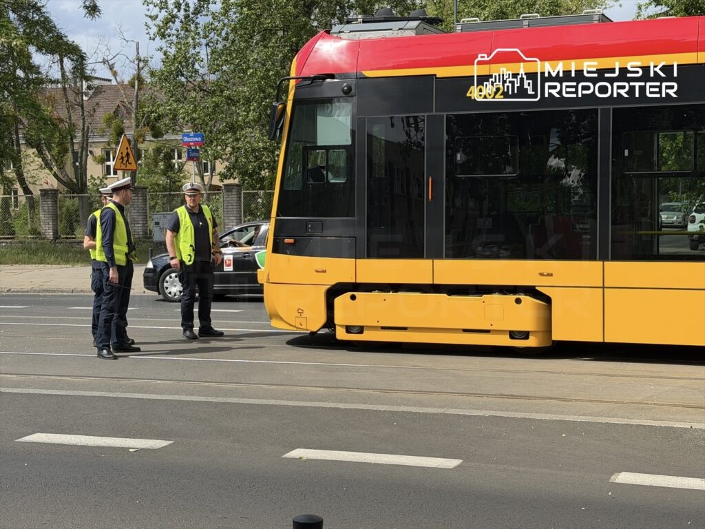 Dwaj mężczyźni w odblaskowych kamizelkach rozmawiają obok żółtego tramwaju zatrzymanego na ulicy.