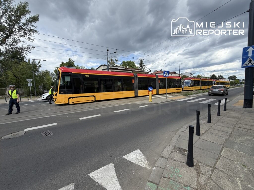 Tramwaj w żółto-czerwonym malowaniu zatrzymany na ulicy, obok dwóch policjantów w kamizelkach odblaskowych.