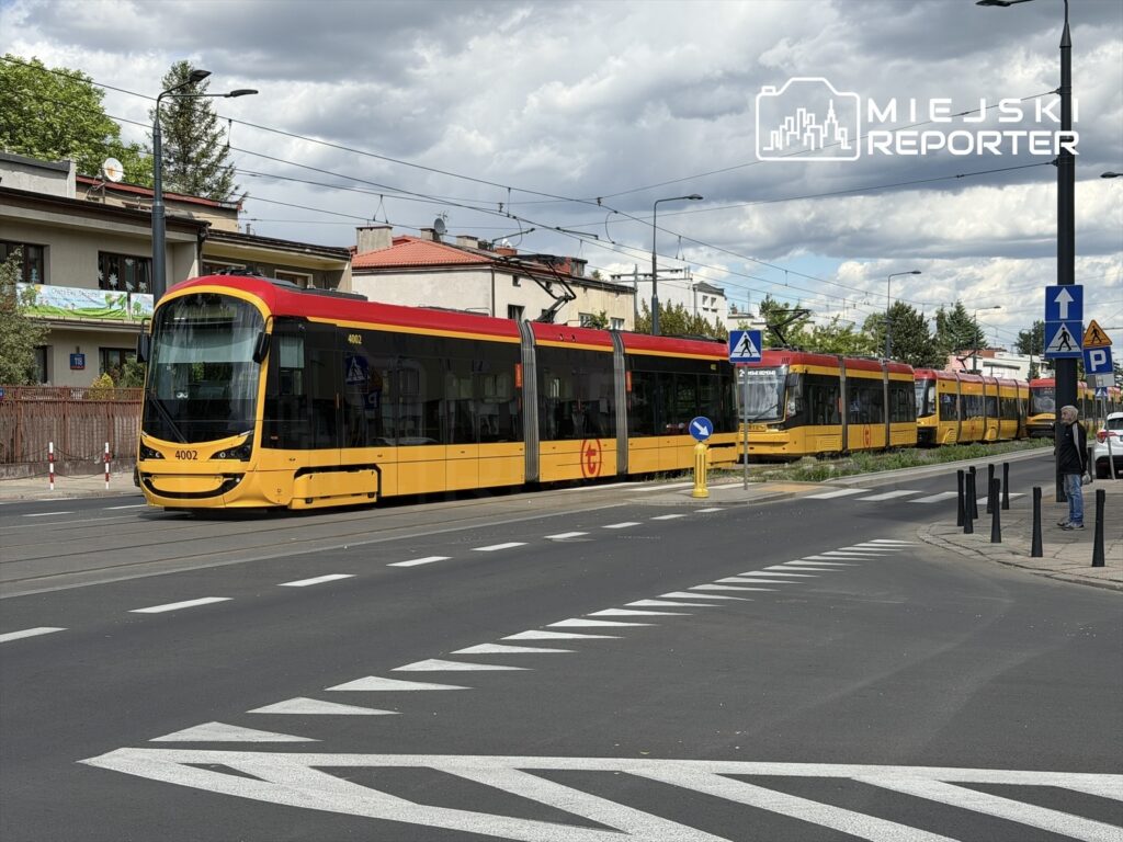 Tramwaj w kolorze żółto-czerwonym zatrzymany na przystanku w miejskim otoczeniu, obok budynków mieszkalnych.