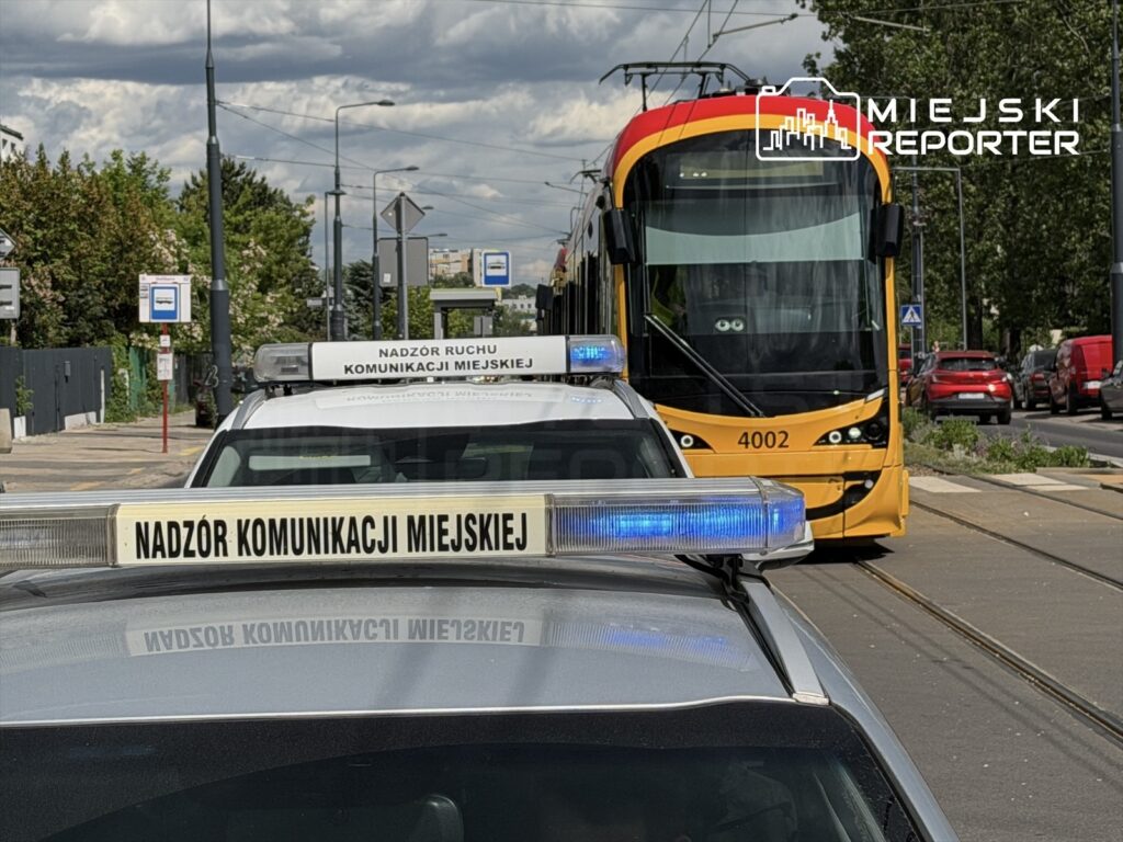 Radiowóz z napisem "Nadzór Ruchu Komunikacji Miejskiej" zatrzymany obok tramwaju w mieście, w tle zieleń i inne pojazdy.