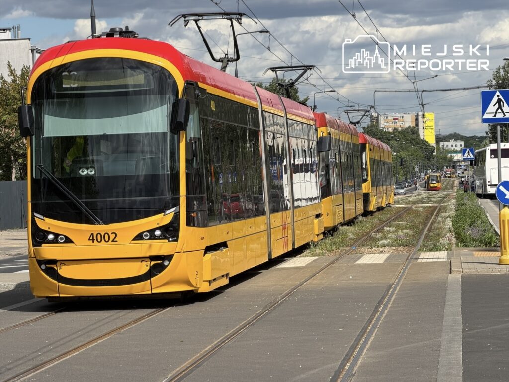 Tramwaj linii 4002 zatrzymany na torowisku w mieście, w tle widoczne budynki i zieleń.