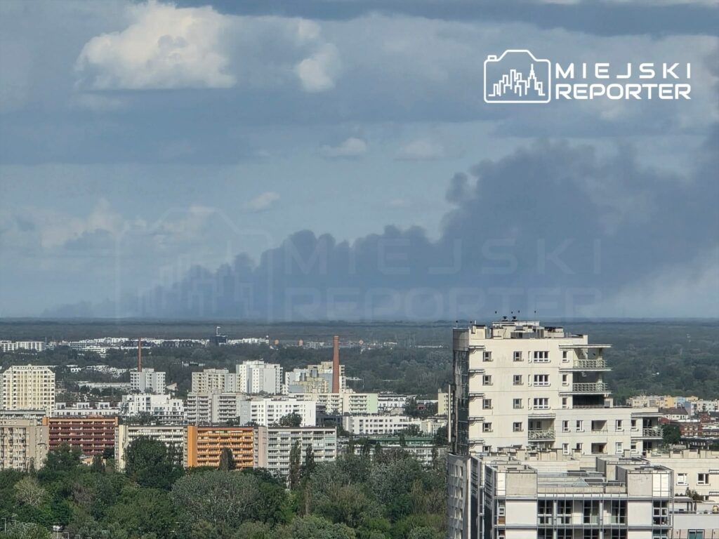 Gęsty dym unoszący się nad miastem, w tle widoczne budynki mieszkalne i zieleń.