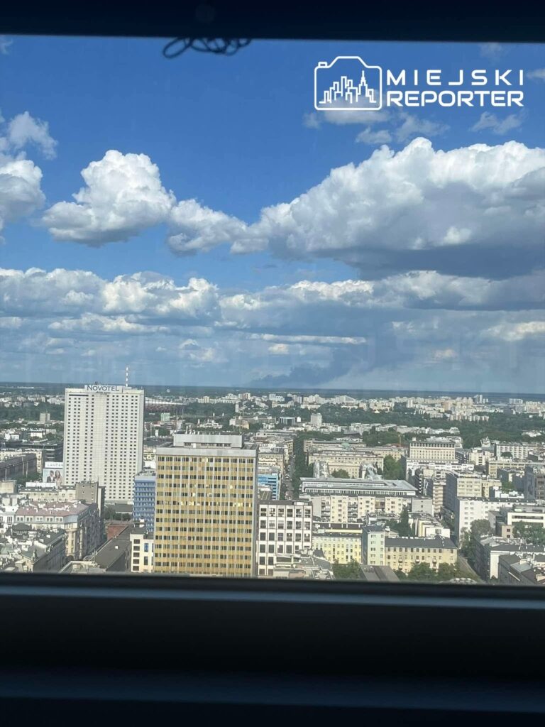 Widok z okna na miejską panoramę z nowoczesnymi budynkami i błękitnym niebem pokrytym białymi chmurami.