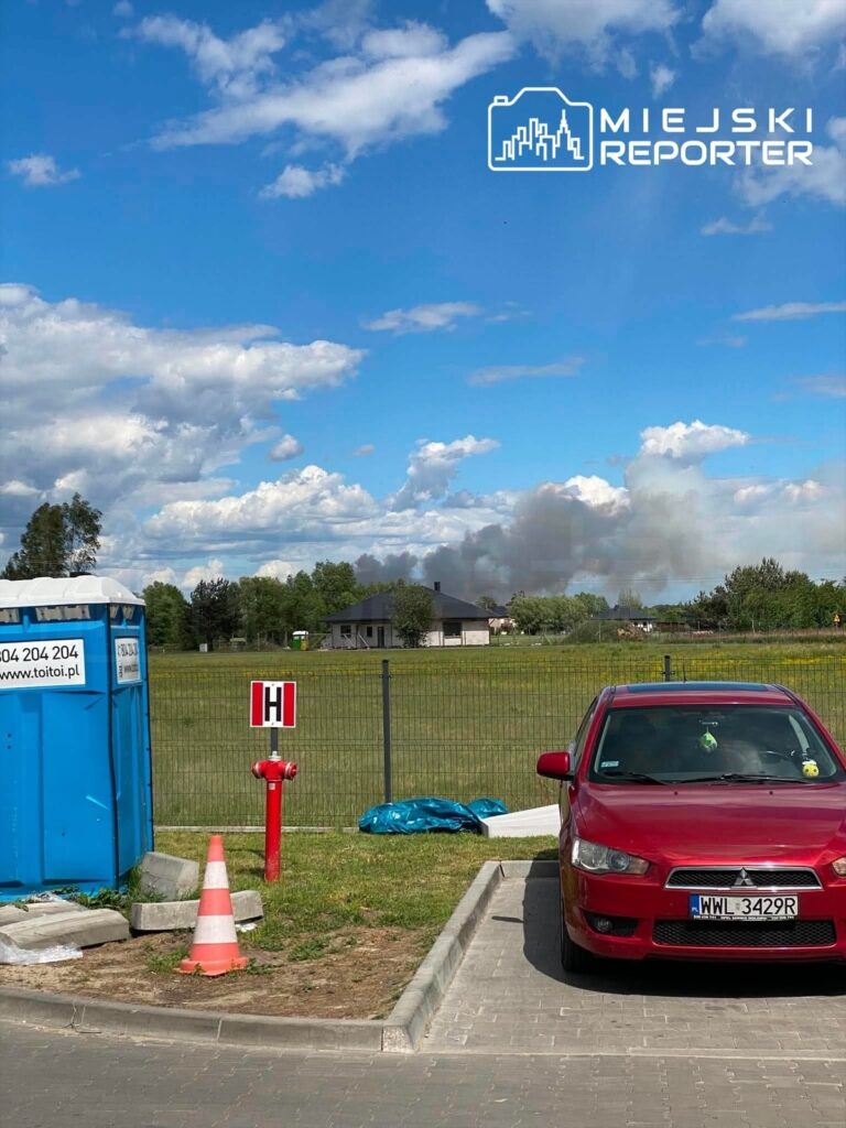 Czerwony samochód osobowy zaparkowany na parkingu obok niebieskiej toalety przenośnej, w tle widoczny dym.