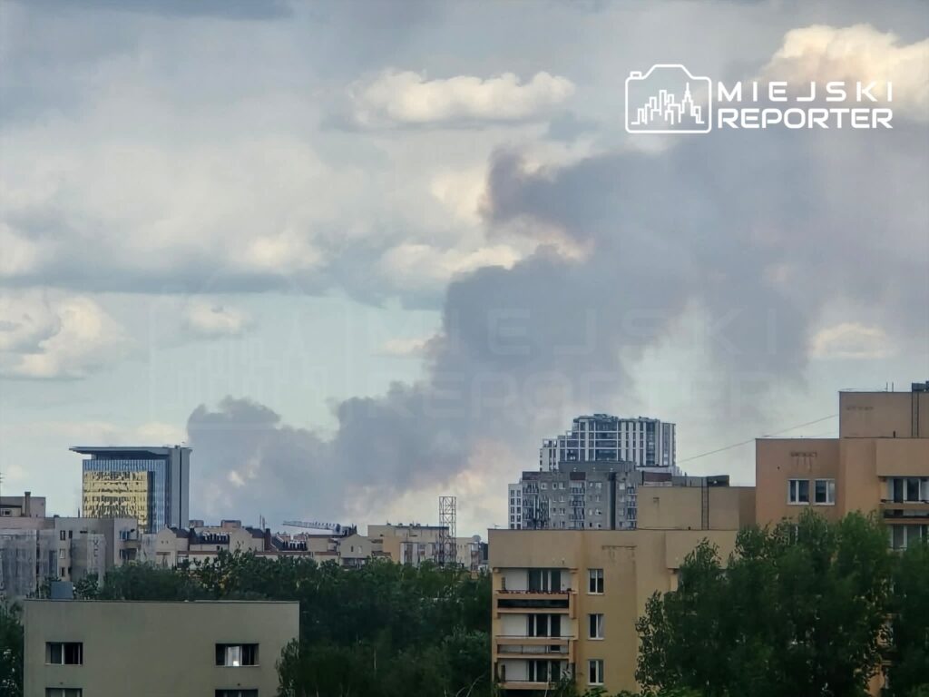 Ciemne chmury dymu unoszą się nad miastem, w tle nowoczesne budynki oraz zieleń drzew.