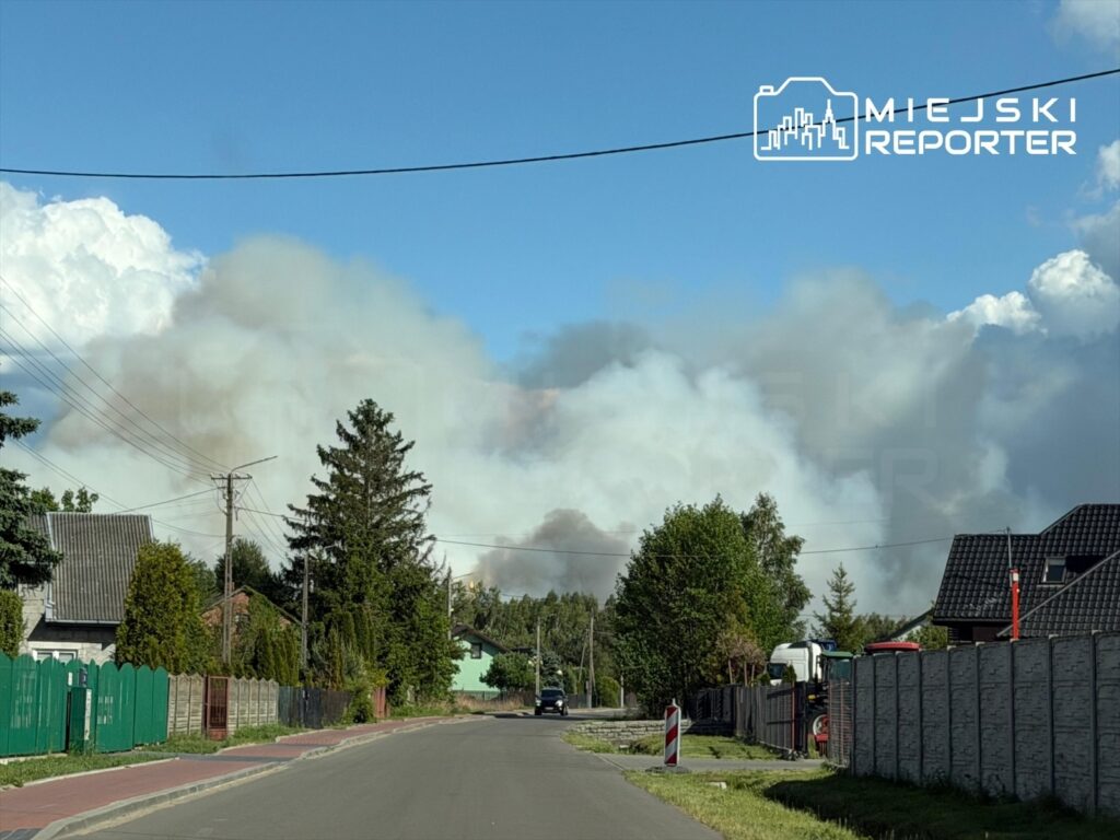 Gęsty dym unoszący się nad zabudowaniami w wiejskiej okolicy, w tle niebieskie niebo z białymi chmurami.