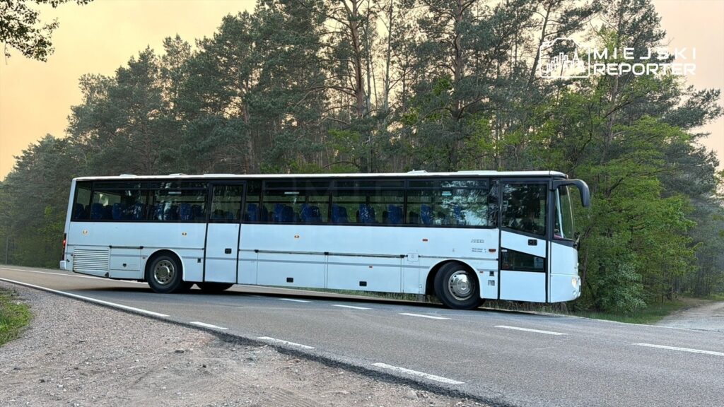 Autobus w białym kolorze zatrzymany na poboczu drogi, otoczony lasem sosnowym.