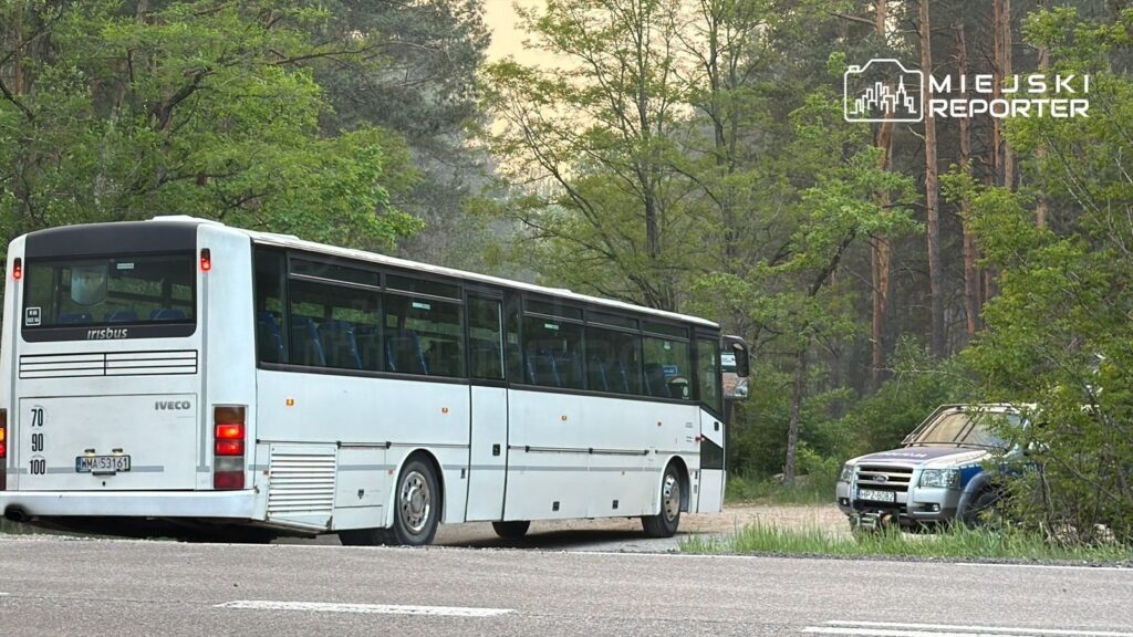 Autobus marki Iveco skręca z drogi na leśną ścieżkę, obok stoi niebieski samochód terenowy.