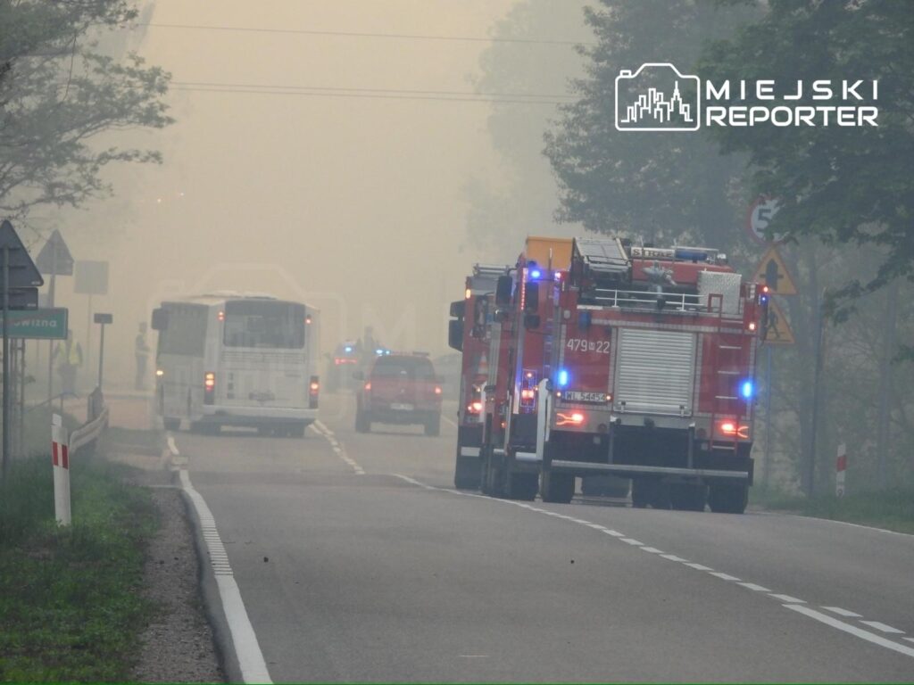 Czerwone wozy strażackie przejeżdżają przez zamgloną drogę, obok białego autobusu i radiowozów policyjnych.