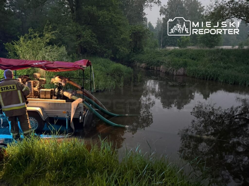 Strażak przy wozie strażackim z pompą, stojący nad rzeką otoczoną zielenią.
