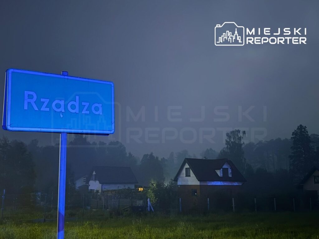 Nieodległy znak drogowy z napisem "Rządz" oświetlony niebieskim światłem, w tle widoczne domy w mglistej scenerii.