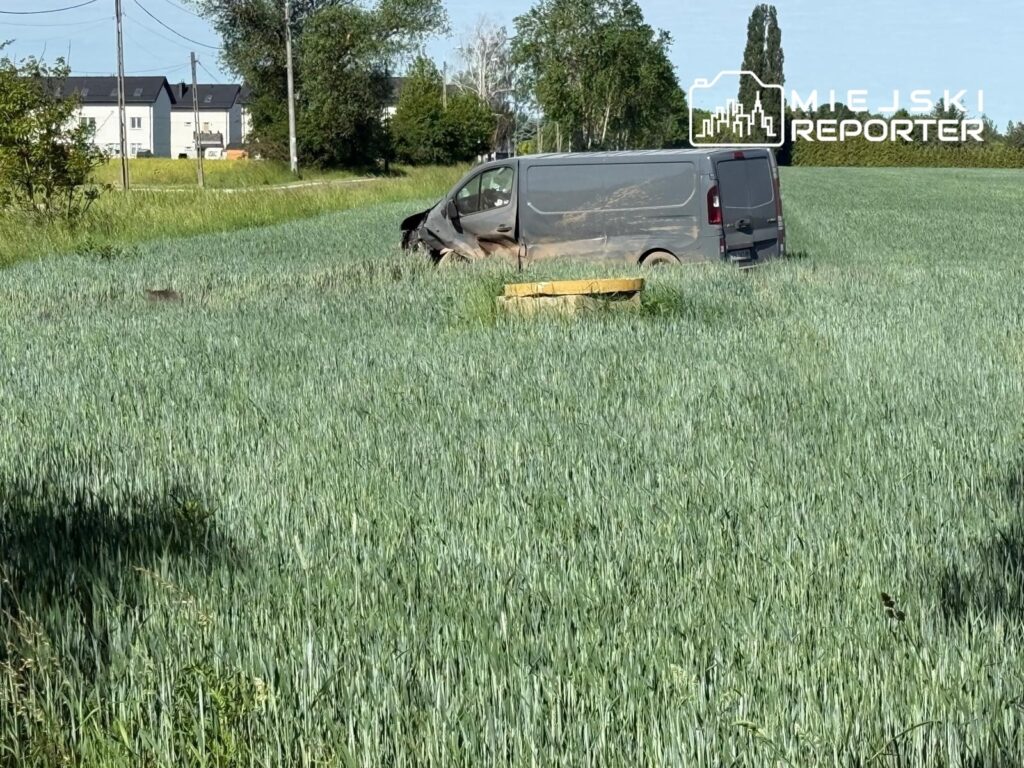Samochód dostawczy utknął w zielonym polu, otoczony wysoką trawą, w tle widoczne budynki mieszkalne.