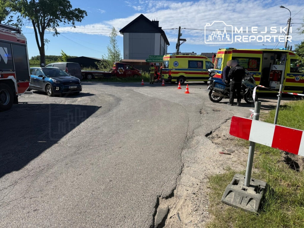 Na skrzyżowaniu ulicy stoją pojazdy straży pożarnej oraz karetki, a policjant rozmawia z osobą na motocyklu.