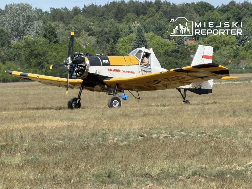 Samolot PZL M-18 Dromader ląduje na polu, w tle widoczne drzewa i zieleń.