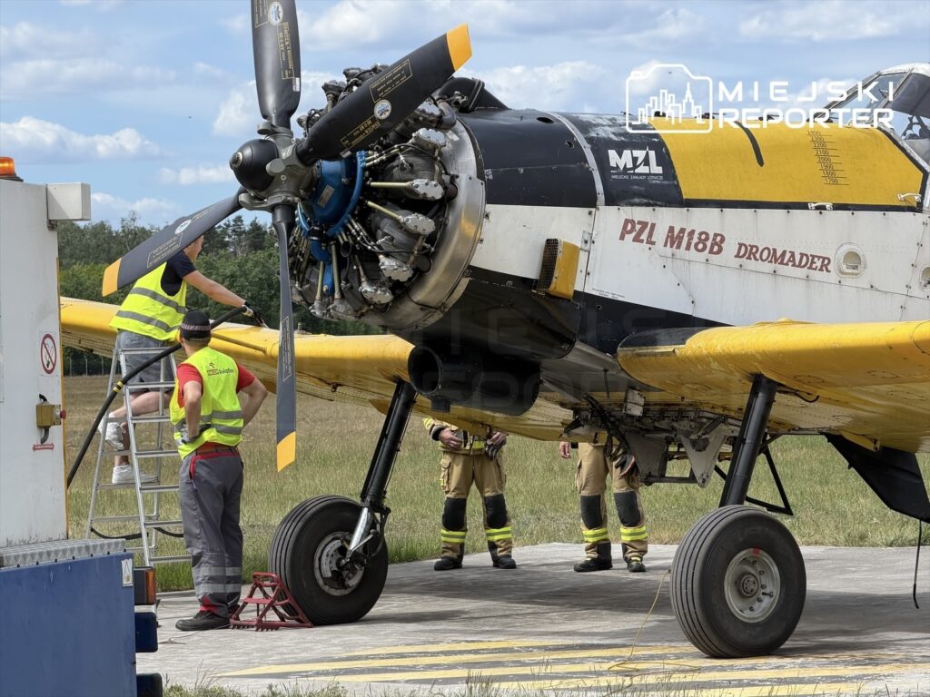 Mężczyźni w kamizelkach odblaskowych pracują przy silniku samolotu PZL M18B Dromader na płycie lotniska.