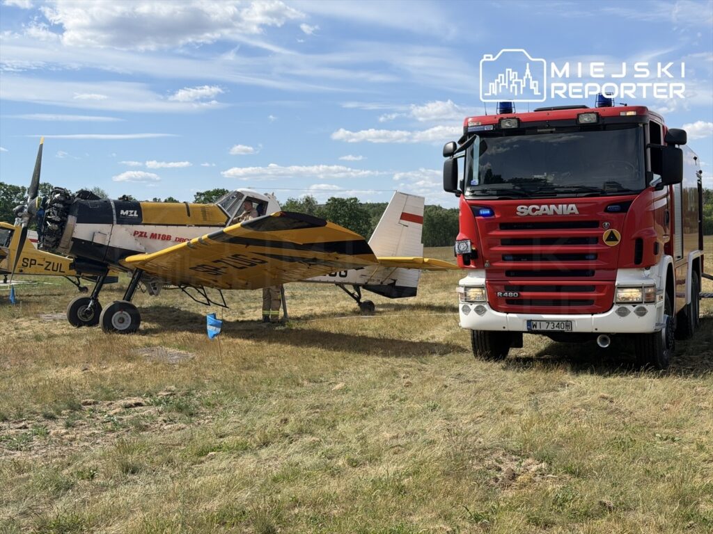 Samolot PZL M18B Dromader stoi na trawiastym terenie obok czerwonego wozu strażackiego Scania.