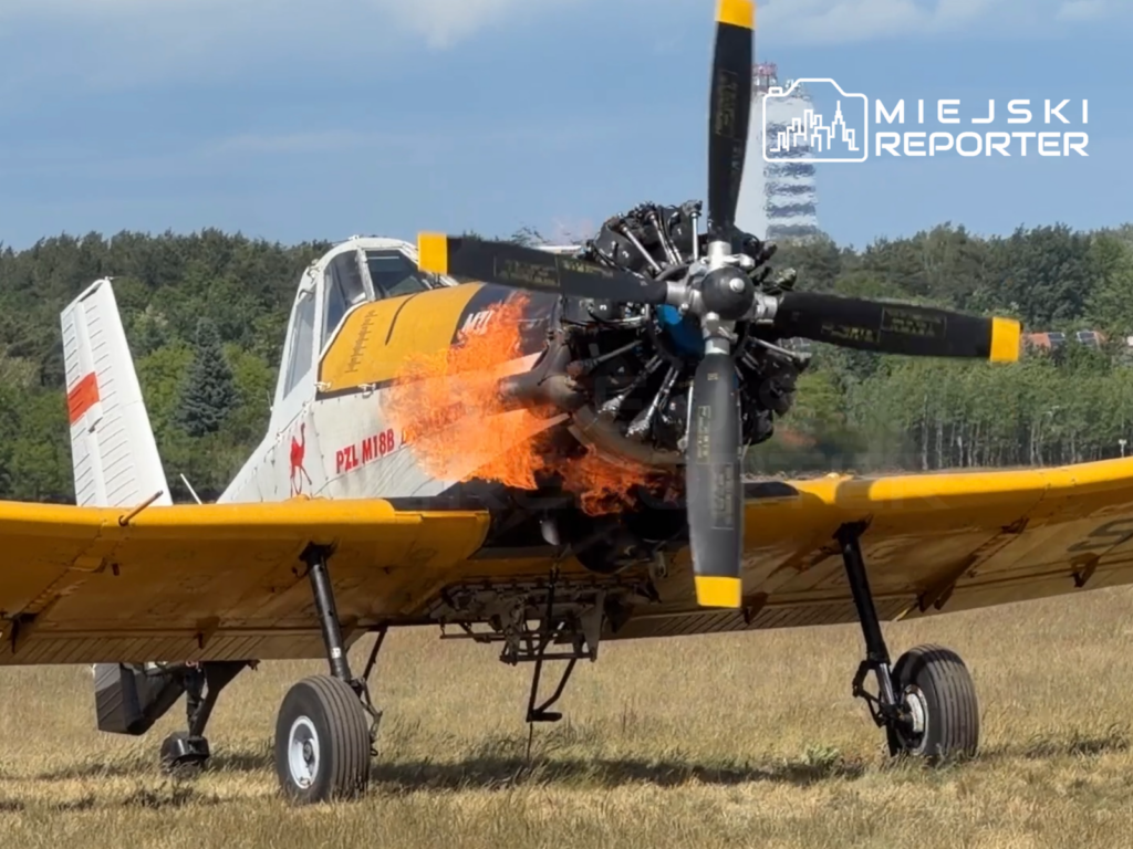 Samolot PZL M188 z płomieniami wydobywającymi się z silnika, stojący na polu z zielonymi drzewami w tle.