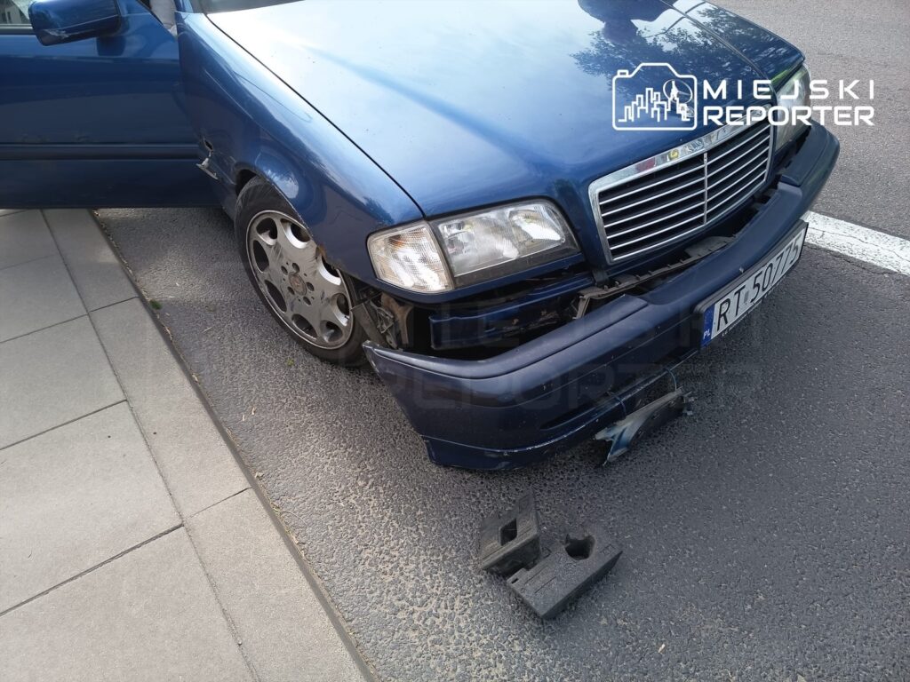 Uszkodzony niebieski samochód osobowy z otwartymi drzwiami zaparkowany na chodniku, obok leżą fragmenty zderzaka.