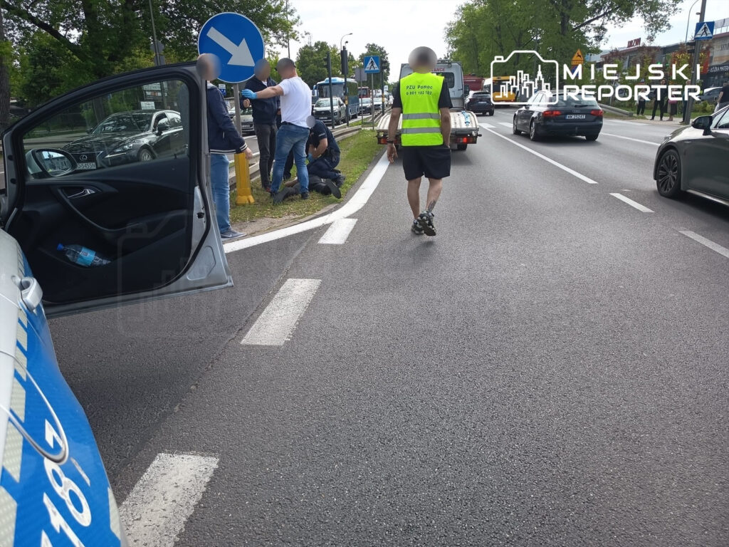Mężczyzna w kamizelce odblaskowej idzie wzdłuż ruchliwej ulicy, obok policjantów interweniujących przy oznakowanym słupku.