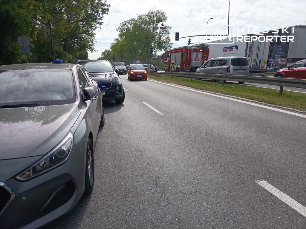 Ruch drogowy z zablokowanym pasem, w tle pojazdy służb ratunkowych i sygnalizacja świetlna.
