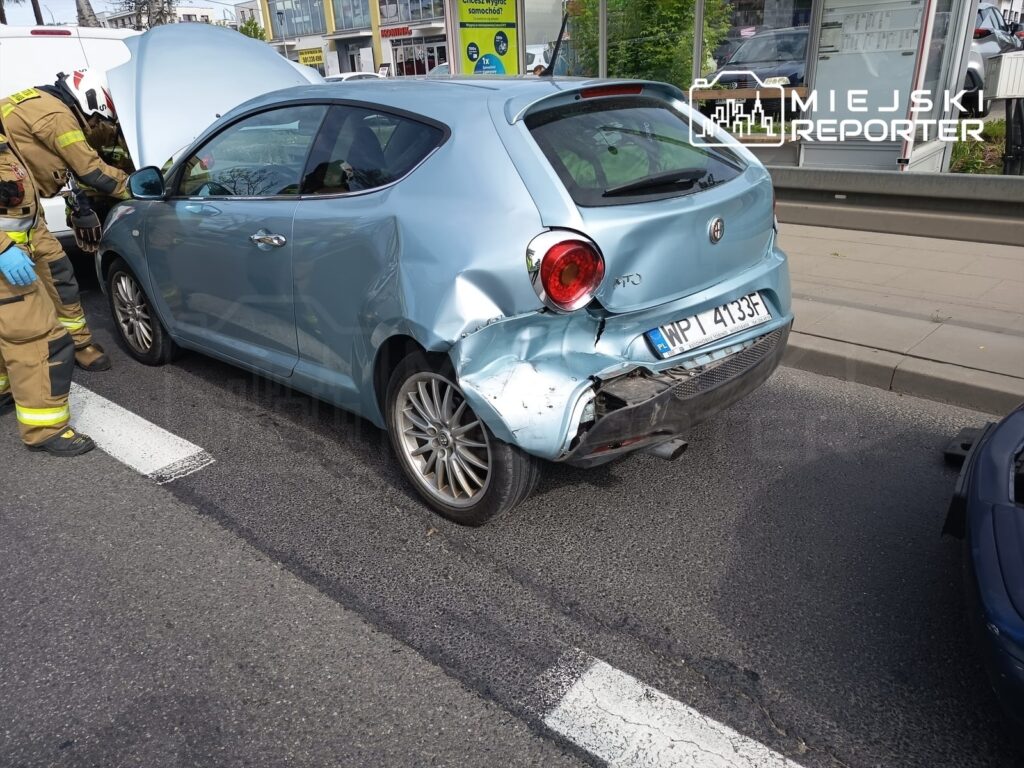Kierowca w mundurze strażackim sprawdza uszkodzone niebieskie auto po kolizji na miejskiej ulicy.