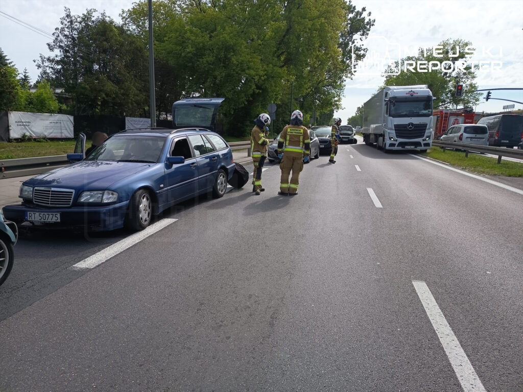 Kierowcy w mundurach strażackich udzielają pomocy przy uszkodzonym niebieskim samochodzie osobowym na drodze.