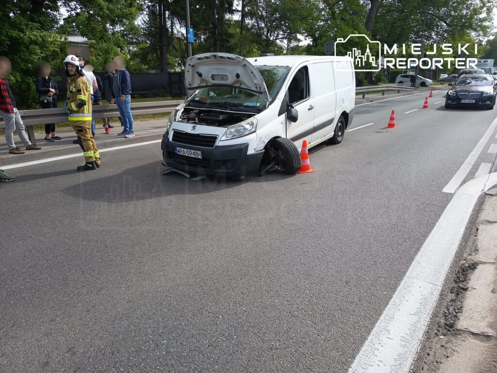 Samochód dostawczy z otwartą maską stoi na drodze, obok niego strażak rozmawia z grupą ludzi, a w tle widoczny ruch uliczny.
