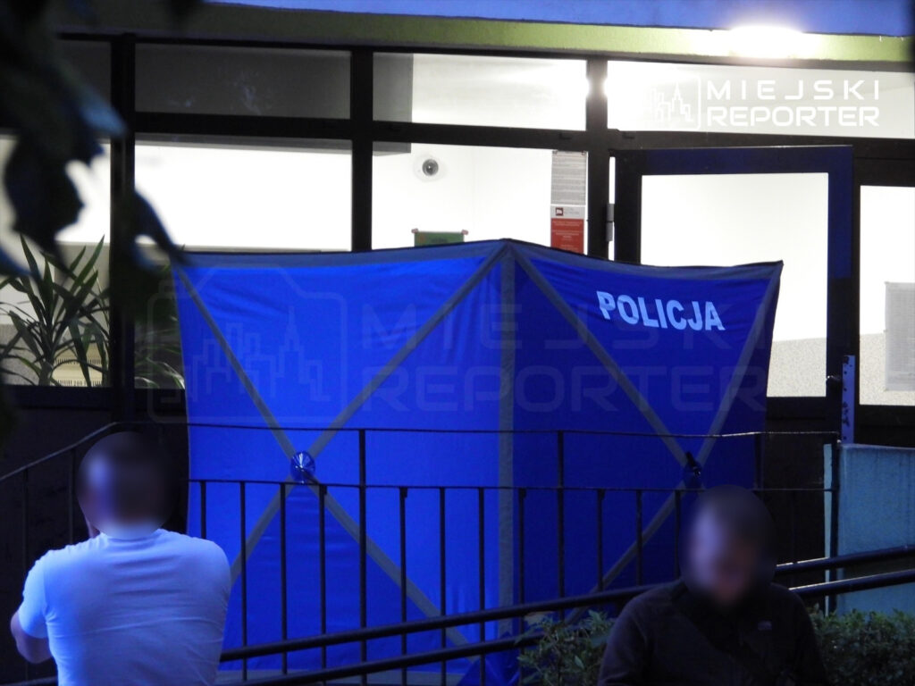 Mężczyzna w białej koszulce siedzi na ławce przed niebieską namiotem policyjnym w nocy.