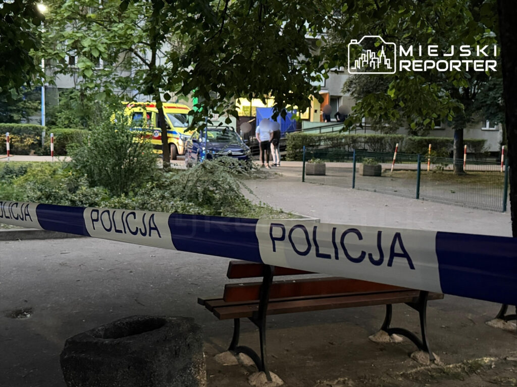 Policjanci zabezpieczają teren z niebieską taśmą w parku, w tle widoczne karetki pogotowia oraz osoby wchodzące do budynku.