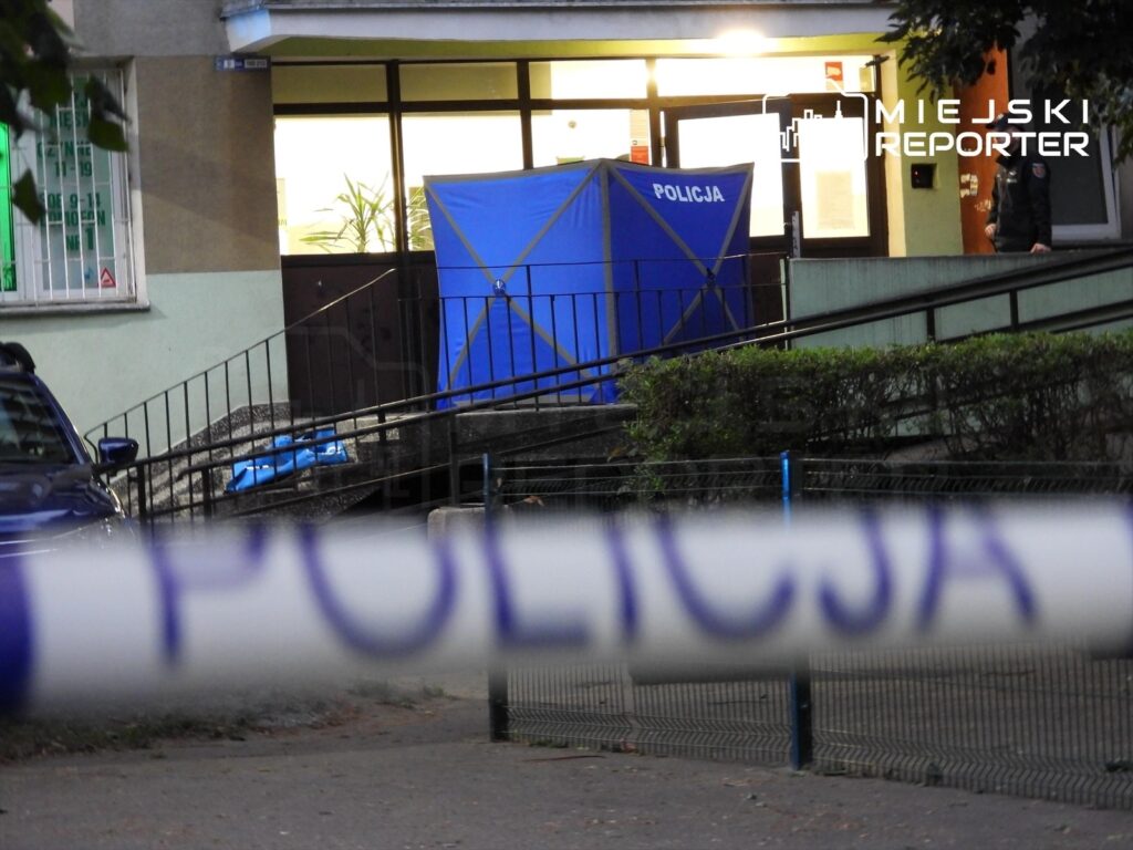 Policjant stoi obok niebieskiego namiotu z napisem "POLICJA" przed wejściem do budynku mieszkalnego.