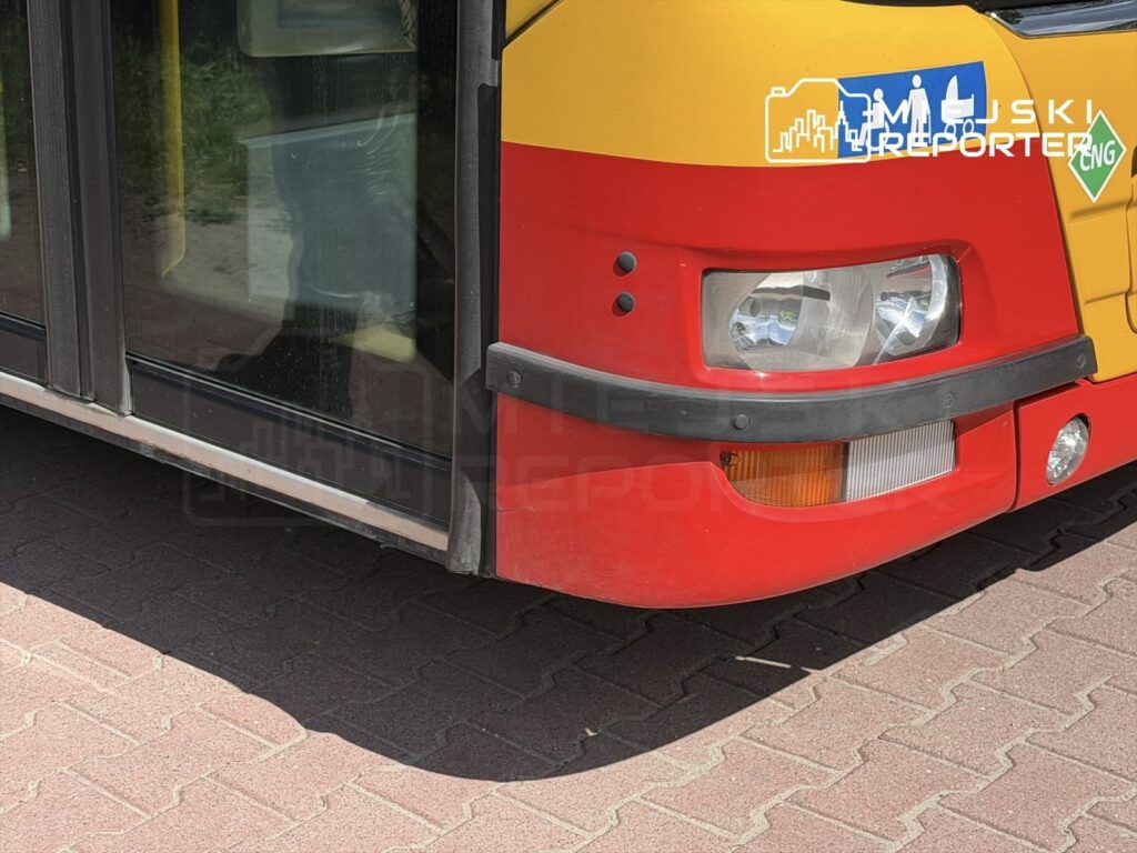 Czerwony autobus z przeszklonymi drzwiami stojący na kostce brukowej, z widocznymi reflektorami i oznaczeniami.