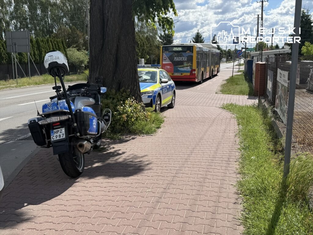 Motocykl policyjny zaparkowany na chodniku obok radiowozu, w tle przejeżdżający autobus na ulicy.