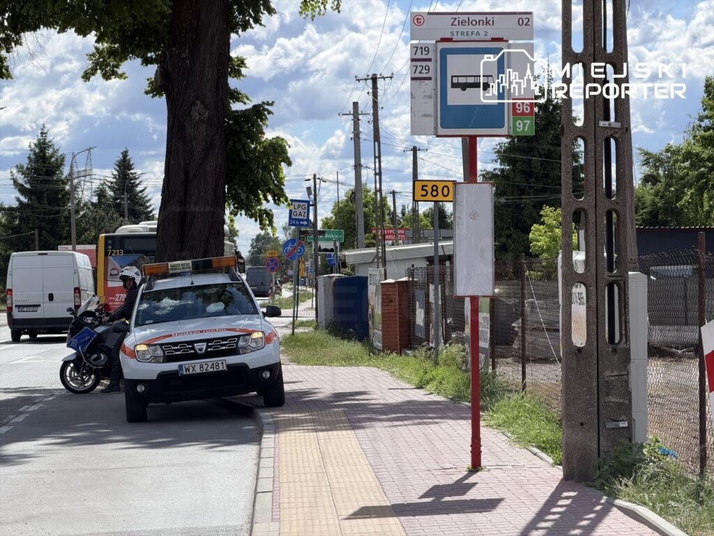 Radiowóz policyjny zaparkowany przy przystanku autobusowym, obok motocykl policyjny, w tle widoczne pojazdy i drzewa.