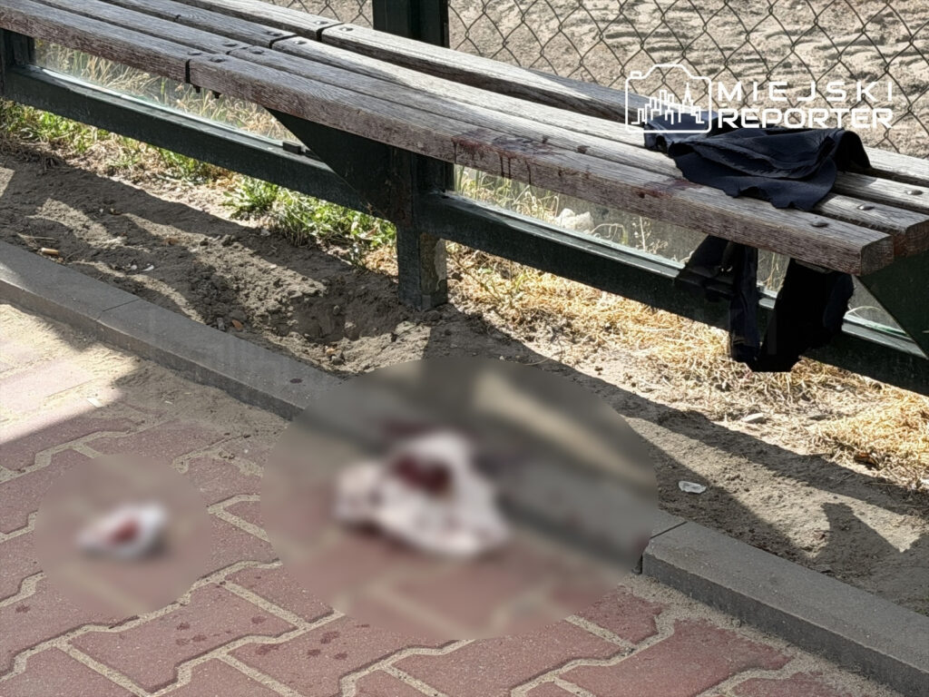 Na ławce w parku leży czarna odzież, a obok na chodniku widoczne są plamy krwi oraz nieczytelne obiekty.